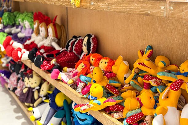 A delightful pile of colorful, handmade stuffed toys, resembling stylized fish and other playful creatures, neatly displayed on a wooden shelf in a market or shop. The vibrant textiles and unique designs showcase the charming folk art and traditional craftsmanship prominent in Bangladesh and South Asia, appealing as gifts or souvenirs.