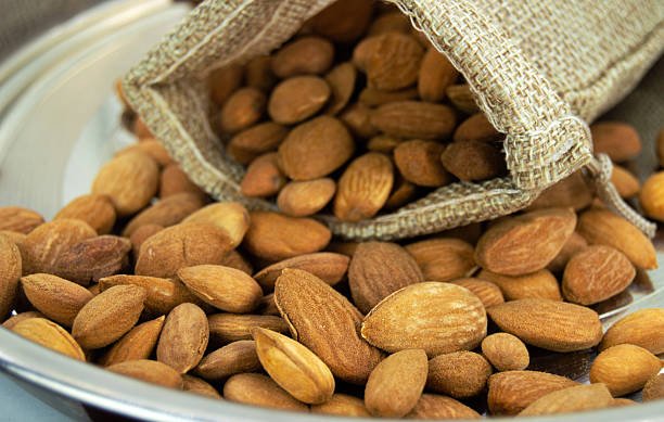 One fallen beige light brown burlap sack gunny bag full of spilling falling snacks overflowing spilled almonds badam healthy nuts in stainless steel plate - table top high angle view. Like prassad, prashaad or religious offering for Diwali, Dussehra, Ganesh Chaturthi.